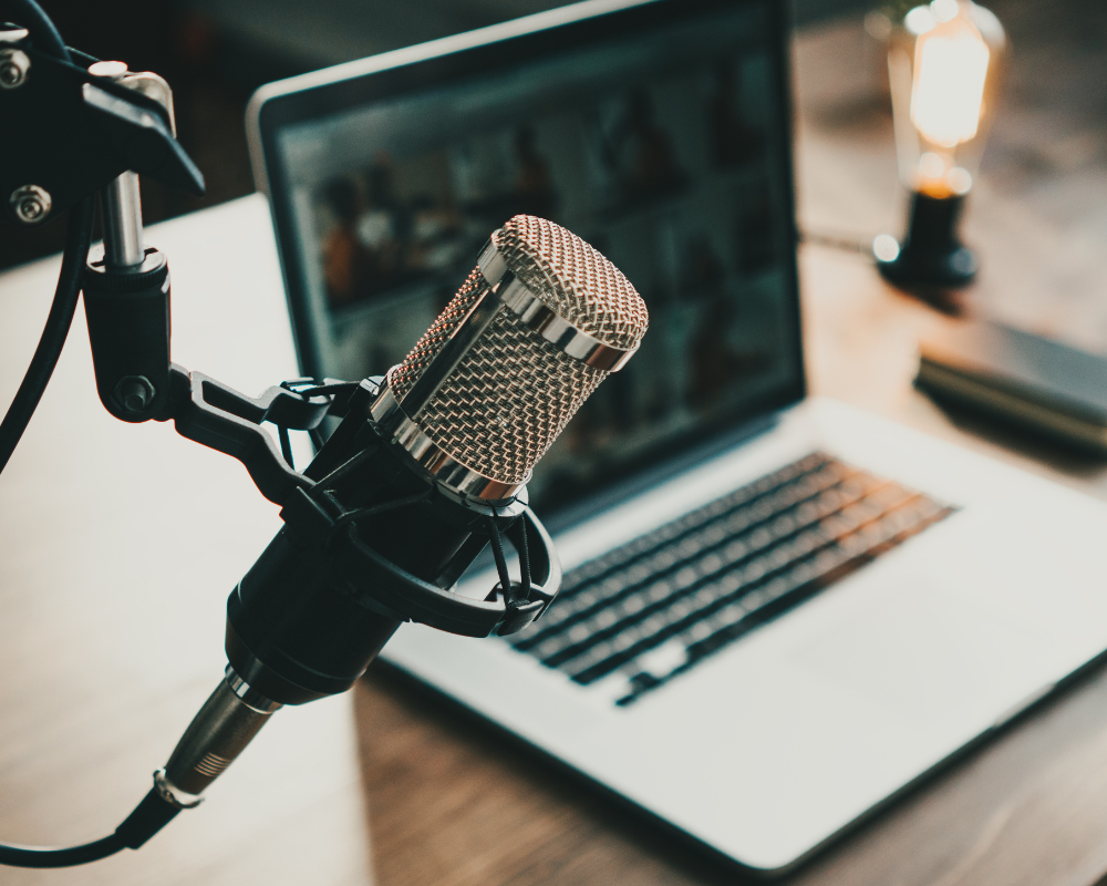 Close-up image of a podcasting setup featuring a mounted microphone placed next to an open laptop. The background shows a lit workspace with a glowing vintage-style bulb and notebooks.