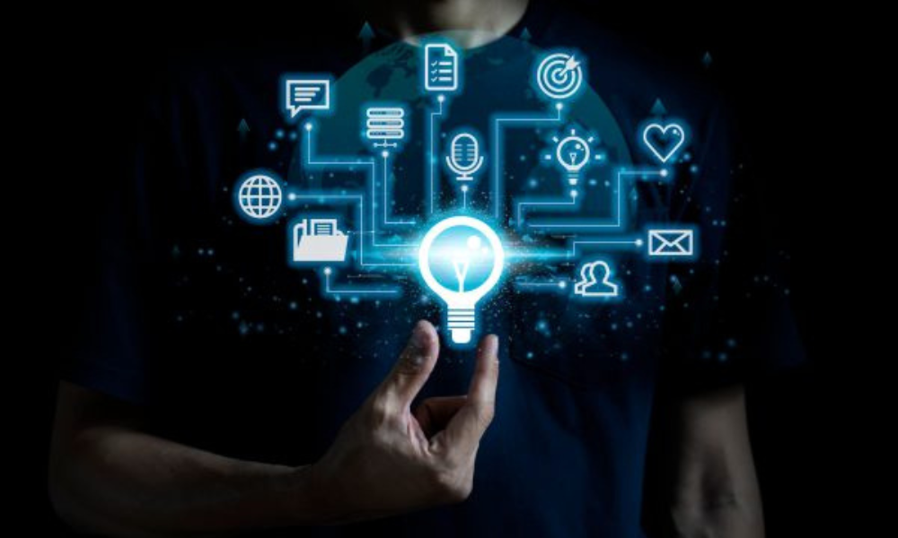 A person, wearing a dark-blue t-shirt, is shown holding a glowing, light-blue digital representation of a lightbulb. The lightbulb appears to be suspended in mid-air. Around the lightbulb are various digital icons, including a globe, a folder, chat bubbles, a microphone, a network icon, a target, a heart shape, and email symbols, all connected by glowing light-blue lines. The background is dark. The overall impression is of a concept of innovation, connectedness, and digital ideas. The person's hands are the focal point, showcasing a conceptual rendering of a connection between various aspects of a modern business or IT environment.