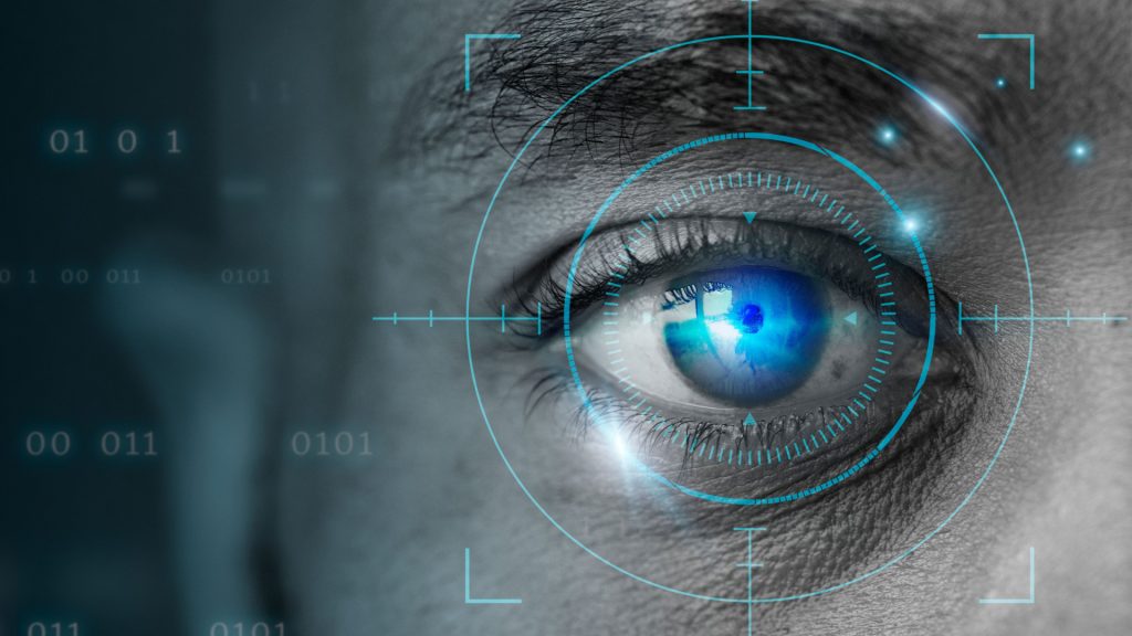 Close-up view of a human eye, likely digitally enhanced. A light teal/cyan circular target-like graphic is superimposed over the eye, with crosshairs. Binary code (0s and 1s) is visible in the background, appearing as a blurry overlay. The eye itself is a deep blue, and the surrounding skin tones are grayscale, appearing somewhat muted or faded. The graphic elements give the impression of biometric scanning or identification technology.