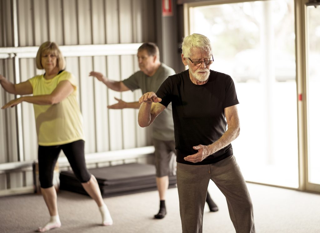 Full indoor shot of several older adults participating in a Tai Chi class. Three adults are visible in the image; the focus is on a man with short grey hair and a grey beard, wearing a dark grey/black t-shirt and light brown/grey pants. He is demonstrating a Tai Chi movement with his arms open, palms facing the viewer. Two other adults, a woman with shoulder-length blonde hair in a yellow top and black pants, and a man with short grey hair in a dark grey/black top and pants are also clearly shown in the image practicing alongside him. They are performing similar poses. The class appears to be taking place in a large, light-coloured, industrial-style building or warehouse with a wide expanse of light tan-grey metal panels for the walls. There is an open doorway in the background, beyond which a blurred outdoor scene is visible. On the floor are mats. The lighting is even and ambient, with a soft, natural quality suggestive of an interior space that is well-lit.