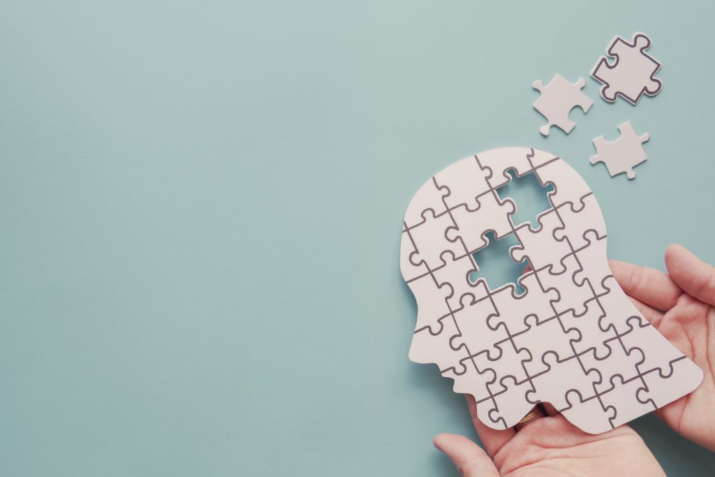 A light teal-blue background. A silhouette of a human head, formed from a large jigsaw puzzle, is centred in the image. The jigsaw pieces are white with dark grey outlines. A section of the puzzle is missing. Some loose jigsaw pieces are scattered around the head silhouette, also white with dark grey outlines. A pair of hands, light-skinned and partially visible, are holding the head silhouette jigsaw.