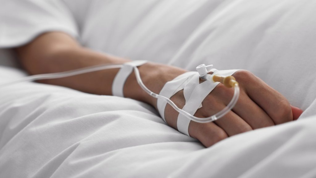 The image depicts a hand with a tube attached, suggesting a medical or therapeutic setting. The scene is set indoors, possibly in a bedroom, where a woman appears to be lying comfortably.