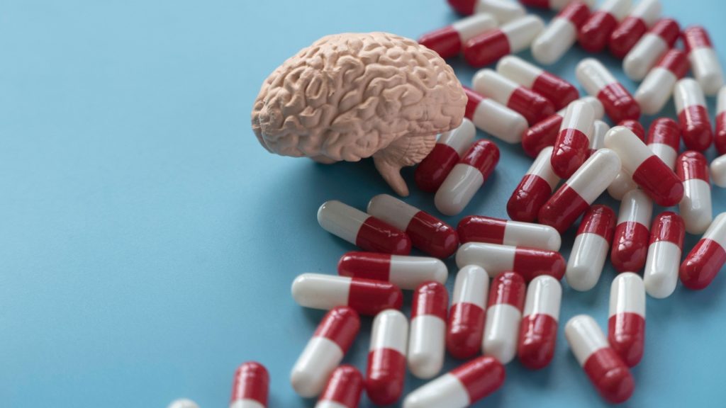 The image features a model of a brain displayed prominently, with various pills arranged around it. The setting suggests a relationship between the brain and pharmaceuticals, emphasizing themes related to medicine and drug use. The color red might indicate urgency or signal importance within the context of health or medication.