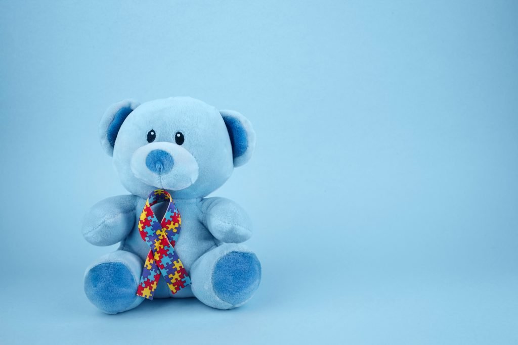 A soft blue teddy bear sits against a matching light blue background. It has a colourful puzzle ribbon around its neck, symbolising autism awareness.